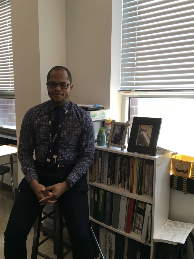 Economics and Personal Finance teacher, Alexander Morton, poses in front of two pictures taken with teachers that have supported him.
